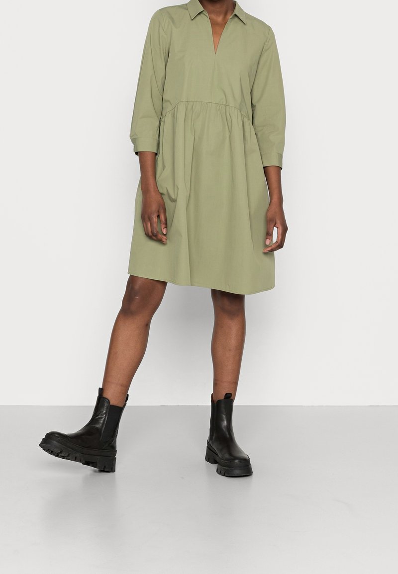 Personne portant une robe ample vert olive, longueur genoux, avec des manches trois-quarts et des bottines noires épaisses, debout sur un sol clair.