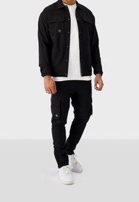 Czarna kurtka z dwoma przednimi kieszeniami i zapięciem na guziki, w zestawie z czarnymi spodniami typu cargo z kieszeniami po bokach, noszona z białymi trampkami.