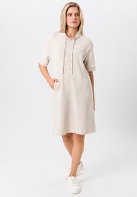 Robe à capuche beige avec des manches courtes, deux poches latérales et un détail de cordon au cou. Tissu lisse, coupe mi-genoux.