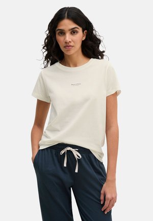 Frau mit langen lockigen Haaren trägt ein weißes T-Shirt mit kurzen Ärmeln und marineblaue Hosen mit Kordelzug, steht mit einer Hand in der Tasche.