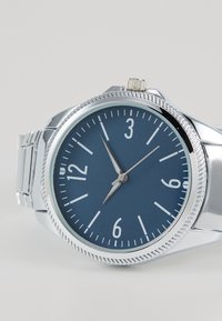 Montre ronde de couleur bleu marine avec un boîtier métallique argenté, index en blanc et une lunette texturée argentée. Présente un design classique et des aiguilles pointues.