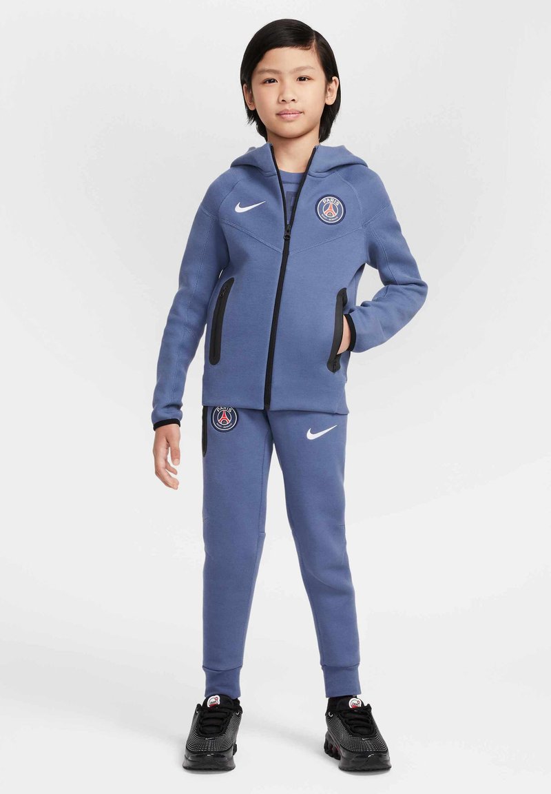 Survêtement Nike bleu avec capuche, fermeture éclair, logo PSG sur la poitrine, accents noirs sur les poches, pantalon slim-fit, porté avec des chaussures de sport noires.
