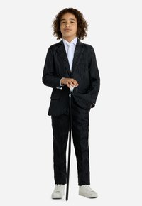 Completo a righe nere con giacca aderente e pantaloni abbinati. Camicia e cravatta bianche, abbinate a sneakers bianche e a un bastone nero.