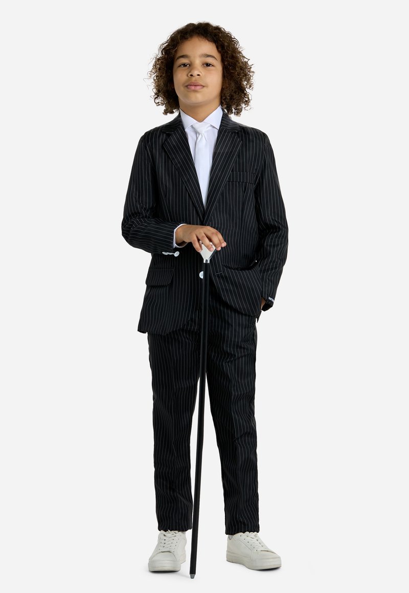 Completo a righe nere con giacca aderente e pantaloni abbinati. Camicia e cravatta bianche, abbinate a sneakers bianche e a un bastone nero.