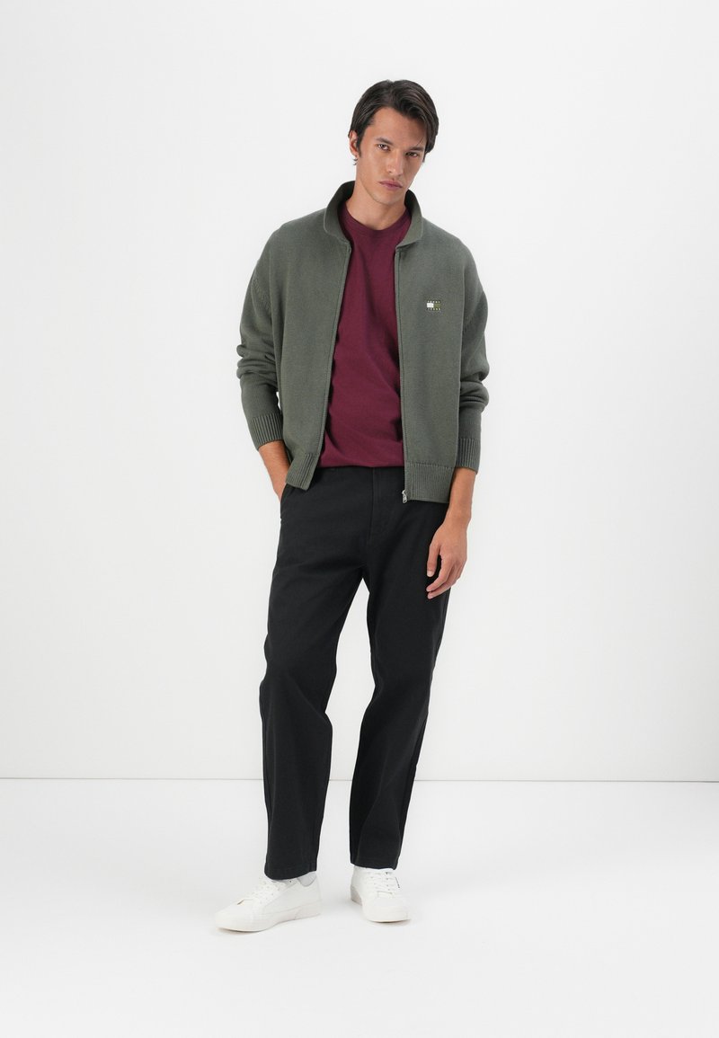 Olive green knitted cardigan with a high collar, paired with a maroon t-shirt and black trousers. White sneakers complete the outfit.