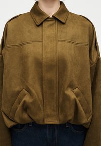 Καφέ σουέντ μπουφάν με μυτερό γιακά, κρυφό μπροστινό κούμπωμα και δύο λοξές τσέπες, φορεμένο πάνω από σκούρα τζιν.