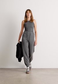 Grå lounge jumpsuit med en tettsittende overdel og løse, avsmalnende ben. Brukt med grå sneakers og holder en svart jakke. Minimalistisk design.