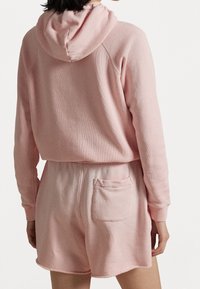 Žena oblečená v měkké růžové mikině a odpovídajících šortkách s zadní kapsou, zobrazená zezadu na jednobarevném b ílém pozadí.