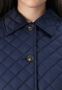 Nærbilde av en kvinnes hals og øvre bryst iført en marineblå quiltet jakke med krage og to knapper, med fokus på teksturen i stoffet.
