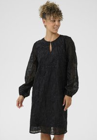 Femeie îmbrăcată cu o rochie neagră până la genunchi, cu mâneci lungi transparente și broderie florală, zâmbind și privind către lateral.