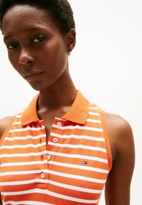 Pomarańczowo-biała koszulka polo w paski z kołnierzykiem, wyposażona w cztery guziki oraz małe logo po lewej stronie. Tkanina teksturowana.