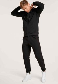 Haut à capuche zippé noir et joggings avec des rayures latérales rouges, fabriqués en matériau doux. Associés à des baskets montantes blanches.