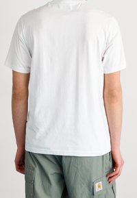 Personne portant un t-shirt blanc uni à manches courtes et un pantalon cargo vert clair avec un patch logo Carhartt visible sur la poche.