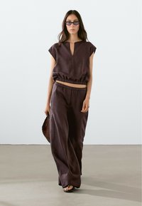 Loose-fitting, dark brown linen outfit with a short-sleeve top, elastic waistband pants, and a textured woven bag.