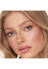 Ein Lipgloss-Applikator mit einer Flockenspitze wird auf volle, glänzende, nudefarbene Lippen aufgetragen. Helle Hautfarbe, mit neutralem Makeup und welligem blonden Haar.