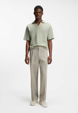 Mann steht und trägt ein hellgrünes kurzärmeliges Hemd, beige hochtaillierte Hosen und beige Sneakers vor weißem Hintergrund.