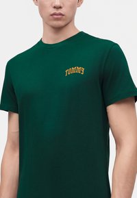Άνδρας που φοράει σκούρο πράσινο t-shirt με κοντά μανίκια και κίτρινο κείμενο "TOMMY" στο αριστερό στήθος, σε απλό γκρι φόντο.