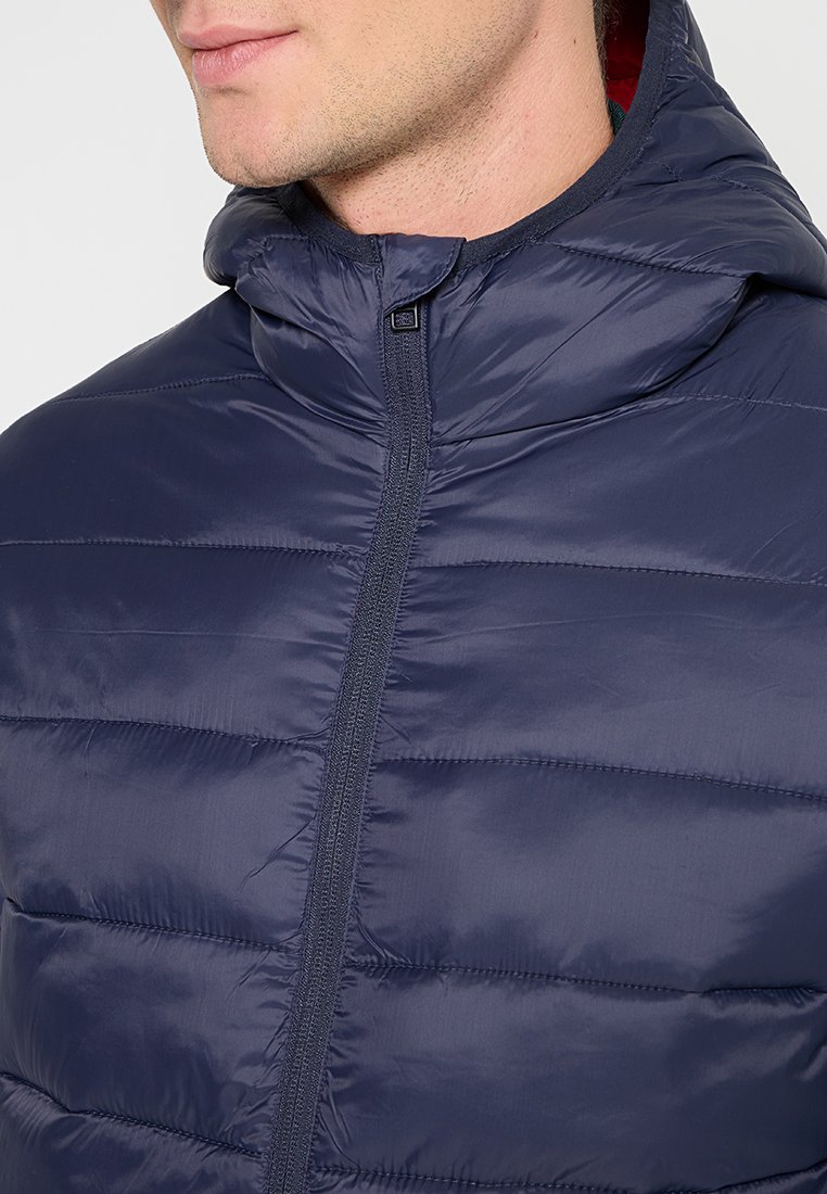 Granatowa kurtka puchowa z wysokim kołnierzem, zamek błyskawiczny, pikowana tekstura i błyszczące wykończenie. Wnętrze ma kontrastową czerwoną podszewkę.
