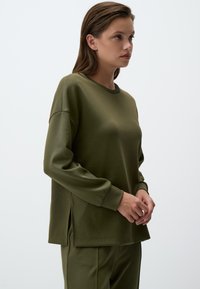 Maglietta a maniche lunghe verde oliva con scollatura rotonda, spalle scese e fessure laterali all'orlo. Tessuto morbido e liscio con una vestibilità rilassata.