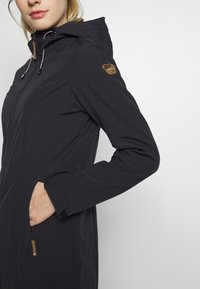 Giacca nera da esterno con zip frontale, cappuccio regolabile e tasche laterali. Presenta un tessuto testurizzato e un piccolo logo sulla spalla.
