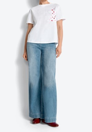 Persoon draagt een wit shirt met korte mouwen met rode hartjes op de zak, lichtblauwe wijde jeans en glanzende rode schoenen, staand tegen een witte muur.