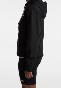 Sudadera negra con un bolsillo tipo canguro, capucha ajustable con cordón y puños de canalé. Combinada con shorts de bicicleta negros con un logo.