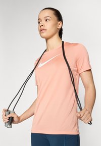 Vaaleanpunainen lyhythihainen urheilupaita, jossa on valkoinen Nike-logomerkki, yhdistettynä hyppynaruun, jossa on mustat langat ja harmaat kahvat.