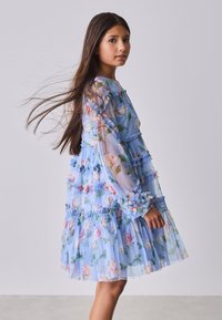 Young girl with long brown hair wearing a light blue floral long-sleeve dress, standing sideways against a plain gray background.