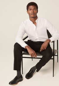 Bijela košulja s ovratnikom, crne uske hlače, crne mokasine, sive čarape. Model sjedi na modernoj crnoj stolici, neutralna pozadina.