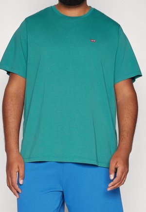 Mand iført teal kortærmet Levi's t-shirt og blå bukser, vist fra nakke til hofter mod en ensfarvet baggrund.