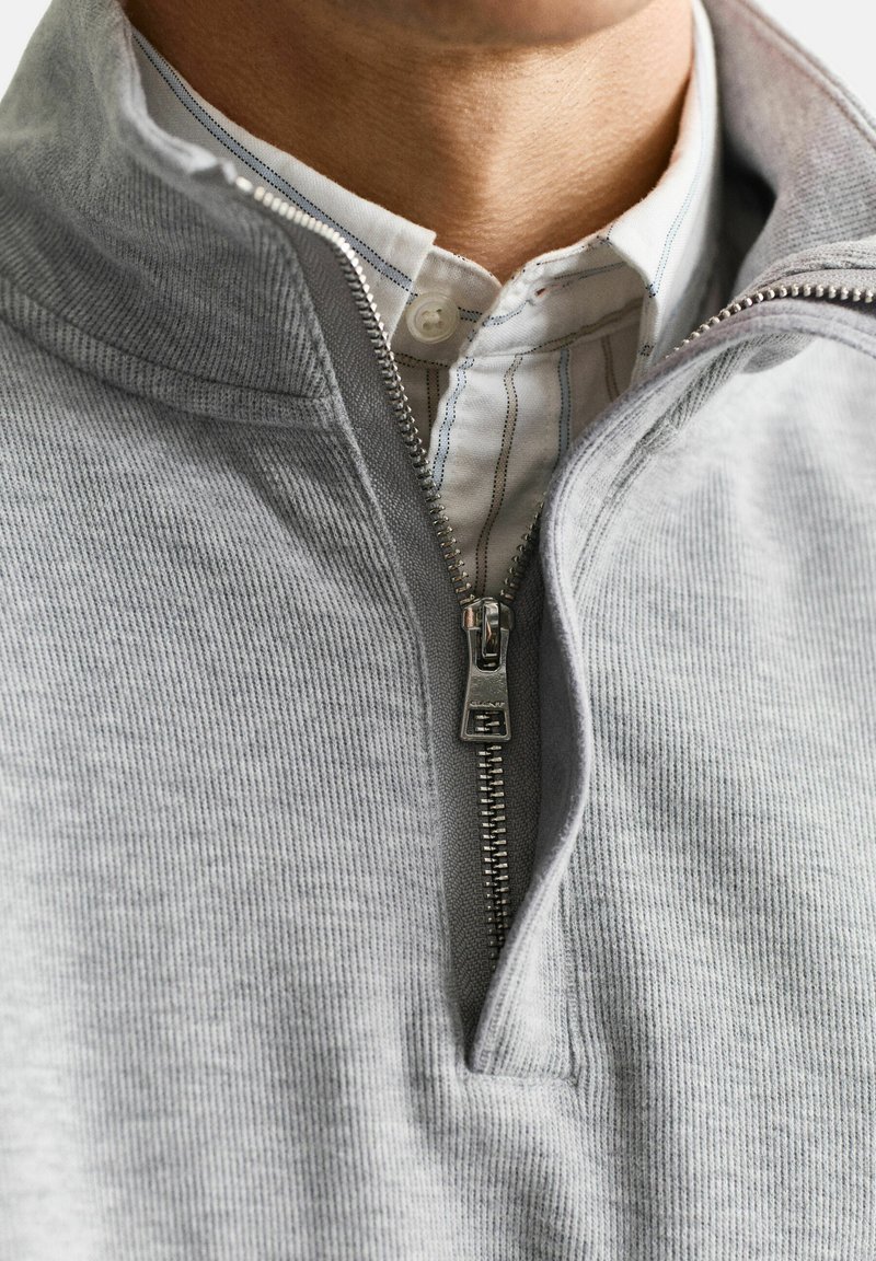 Mann trägt ein weißes Hemd unter einem grauen Pullover mit Viertelreißverschluss, der teilweise geöffnet ist und Kragen sowie Hals zeigt.