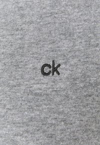 Grå bomuldsstof med en fin tekstur, der har et lille sort broderet "ck" logo. Simpelt design, nærbillede af materialet.