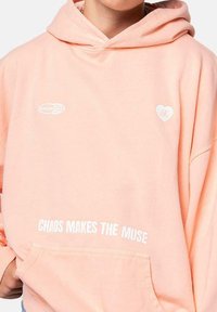 Sudadera con capucha color durazno, que presenta el texto blanco "EL CAOS HACE A LA MUSA" y un pequeño logo de corazón. Tejido suave, corte relajado, bolsillo frontal.