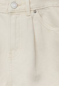 Gros plan d’un jean en denim couleur crème montrant un bouton métallique, un rivet, un passant de ceinture, des coutures et une partie de la poche avant.
