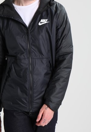 Persoon die een zwart Nike-jas met rits draagt over een wit shirt, met zichtbare tatoeages op de linkerhand en vingers.