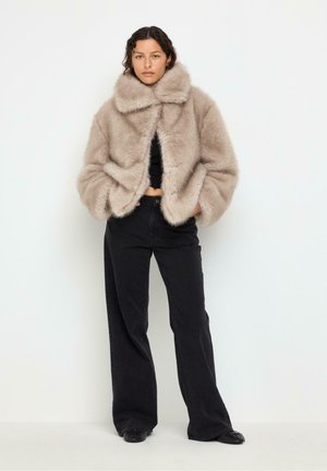 Donna in piedi che indossa una giacca di pelliccia ecologica beige sopra un crop top nero, abbinata a pantaloni neri a gamba larga e scarpe nere su uno sfondo semplice.