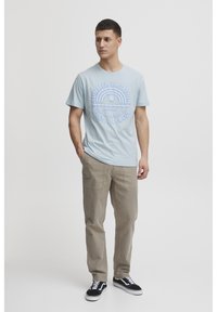 Lyseblå bomull T-skjorte med et rundt grafisk motiv med tekst, kombinert med lysebeige cargo-bukser og svarte joggesko med hvite detaljer.