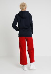 Mornarica dukserica s kenguruvim džepom i velikom kapuljačom, uparen s širokim crvenim samtonim hlačama. Bijele tenisice upotpunjuju outfit.