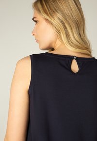 Marineblauwe mouwloze top met een kleine, ronde sleutelgatopening aan de achterkant, accentueert door een knoop. Glad, zacht stoftextuur.