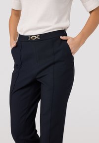 Marineblauwe tailored broek met een zachte textuur, voorzien van een gouden hardware detail op de tailleband en de zijzakken.