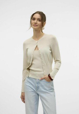 Cárdigan beige con escote redondo y parte delantera de botones, combinado con una camiseta sin mangas a juego. Jeans azul claro, de textura suave y diseño ajustado.