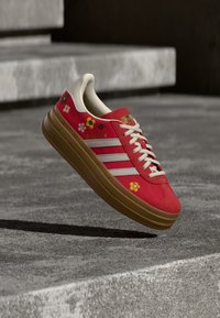 Červená teniska z veluru s květinovou výšivkou, třemi bílými pruhy, gumovou podrážkou a texturovanými zlatými detaily. Zavazování na tkaničky.