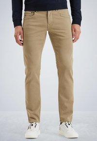 Chino beige in cotone con vestibilità slim e design a cinque tasche, completati da accessori neri e una texture liscia. Indossati con sneakers bianche.