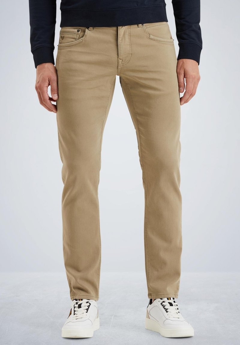 Chino beige in cotone con vestibilità slim e design a cinque tasche, completati da accessori neri e una texture liscia. Indossati con sneakers bianche.