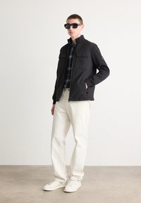 Jachetă neagră cu fermoar, cu două buzunare frontale, purtată peste o cămașă cu carouri. Asortată cu pantaloni de culoare deschisă și adidași albi. Ochelari de soare închiși la culoare.