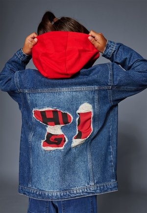 Osoba w dżinsowej kurtce z podartym tyłem, pod którym widać czerwony materiał z czarnymi literami, poprawiająca na głowie czerwoną kapturę.