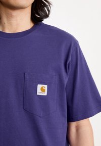 T-shirt in cotone viola con tasca sul petto e un'etichetta quadrata bianca che mostra un logo arancione. Maniche corte, scollatura a girocollo.