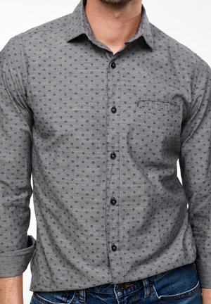 Mann trägt ein graues Hemd mit Knöpfen, das ein dezentes schwarzes Blumenmuster aufweist und mit hochgekrempelten Ärmeln zu blauen Jeans kombiniert ist.