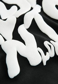 Černá tkanina s texturovaným bílým písmem v zvlněném stylu, obsahující kombinaci silných a tenkých tvarů, vytvářející odvážný vizuální efekt.