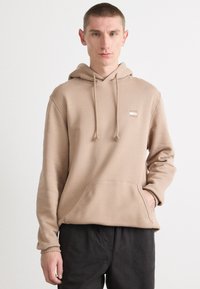 Beige hoodie van zacht materiaal, met een kangoeroezak, capuchon met trekkoord en een klein logo-detail op de borst.